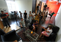 A group of people standing and sitting in a spacious room with dark furniture and a red wall.