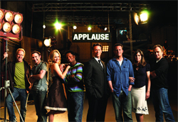 Eight actors posing in a row on a dark television studio set with bright stage lights and an 'APPLAUSE' sign.
