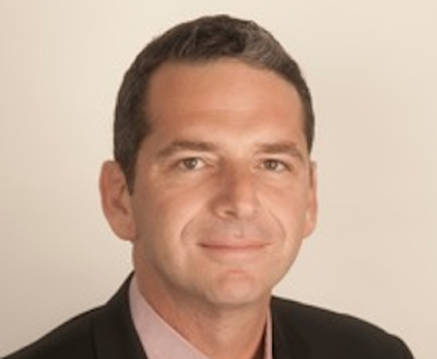 A man with short dark hair wearing a dark suit jacket and a light pink shirt against a plain background.