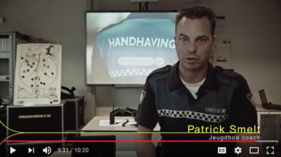 A man in a dark blue uniform with a checkered pattern sits in an office in front of a screen.