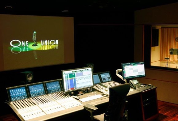 A recording studio control room featuring a large mixing console, multiple computer monitors, and a projection screen.