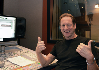 Smiling man in a black t-shirt giving two thumbs up in a recording studio next to a mixing console.