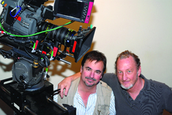 Two men smiling next to a large, complex professional motion picture camera on a mounting rig.