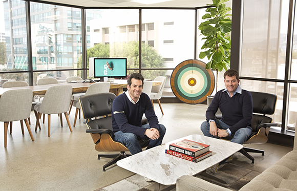 Two men sitting in black leather lounge chairs in a bright, modern office with large windows.
