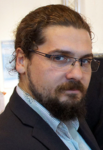 A man with long brown hair tied back, wearing glasses and a dark suit jacket.