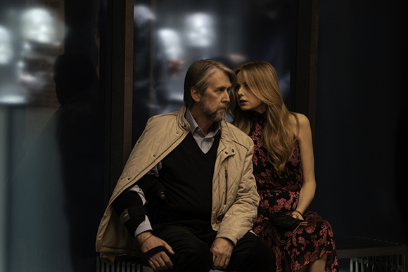 A man in a beige jacket and a woman in a floral dress sit close together in low light.