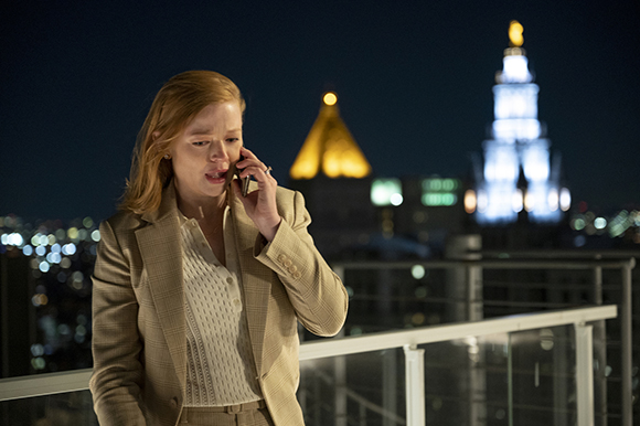 A woman with reddish hair in a tan blazer talks on a cell phone on a balcony overlooking a city at night.