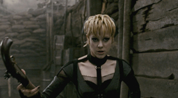 A blonde woman in black attire holds a dark, curved object in a dim, industrial setting.