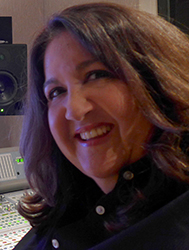 A smiling woman with wavy brown hair wearing a black button-down shirt in a studio.