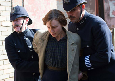Two police officers in dark uniforms and helmets escort a woman in a tan coat and dark blouse.