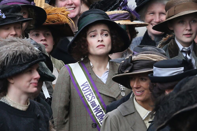 A woman wearing a black hat and a purple and white sash that reads 'Votes for Women' stands among a crowd.