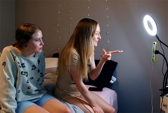 Two young women sit on a bed, looking toward a bright ring light on a stand.