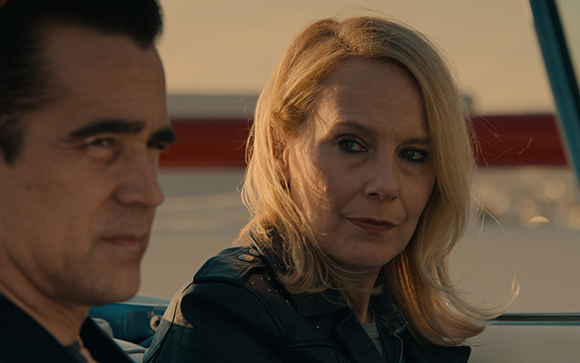 A man with dark hair looks forward while a blonde woman in a leather jacket looks toward him.