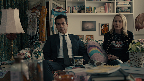 A man in a dark suit and a woman in a black graphic t-shirt sit on a patterned sofa in a room.