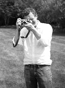 A man in a white shirt and jeans holds a camera to his eye while standing in a grassy field.