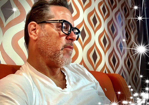 A man with short hair and glasses wearing a white t-shirt against a patterned background.