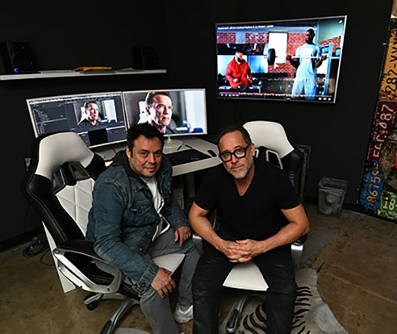 Two men sit in white gaming chairs in a dark studio with several computer monitors displaying video.