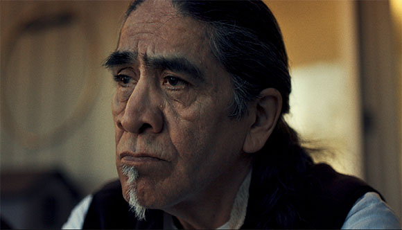Close-up portrait of an older man with long dark hair and a small white goatee looking off-camera.