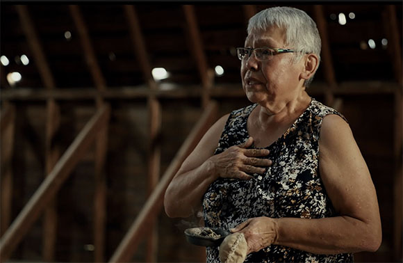 An elderly woman with short grey hair and glasses holds a small bowl and a cloth to her chest.