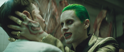 A man with bright green hair and pale skin holds a man's face with a hand featuring a mouth tattoo.