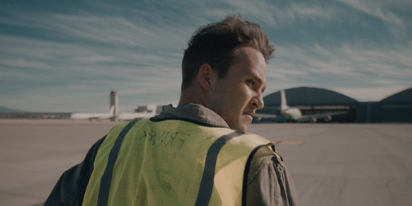 A man wearing a yellow safety vest looks back over his shoulder at an airport tarmac with planes and hangars.