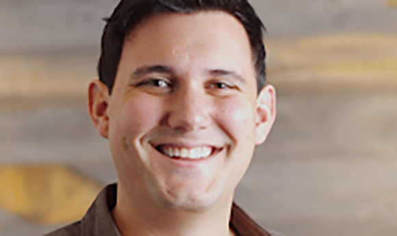 A man with short dark hair smiling broadly against a blurred neutral background.