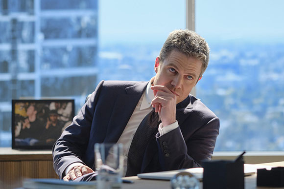 A man in a dark suit and tie rests his chin on his hand while sitting at a desk in a high-rise office.