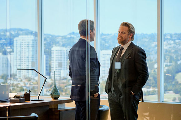 Two men in dark business suits stand in a modern office with large windows overlooking a cityscape.