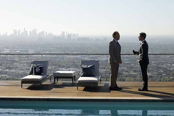 Two men in business suits stand talking on a wooden rooftop deck overlooking a hazy city skyline.