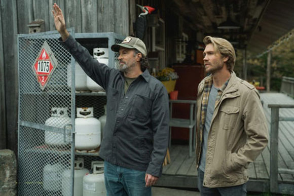 Two men stand outdoors near a metal cage filled with white propane tanks.