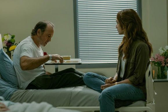 A man in a white t-shirt sits in a hospital bed talking to a woman sitting on the edge of the bed.