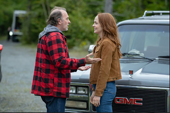 A man in a red plaid jacket and a woman in a brown jacket stand talking next to a grey GMC truck.