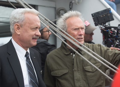 A man in a suit and a man in an olive jacket stand near ropes and a film camera on a boat.