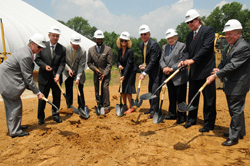 A group of people in business attire and white hard hats hold shovels over a dirt mound during a ceremony.