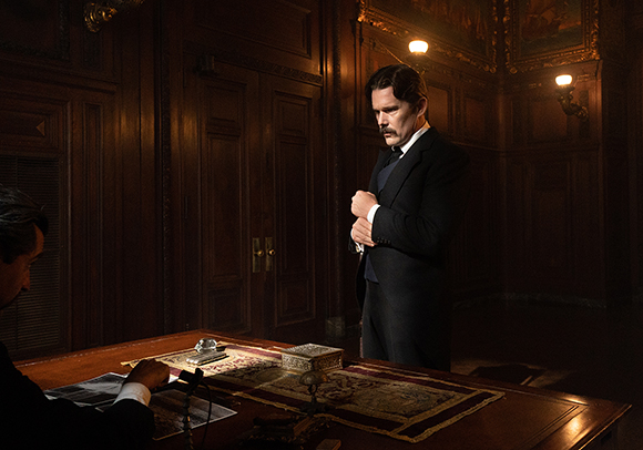 A man in a dark suit and waistcoat stands in a dimly lit, wood-paneled room beside a large desk.