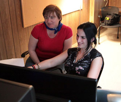 A woman in a red shirt and blue bandana watches a woman in a black patterned top use a computer.