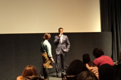 A man in a suit and a woman stand on a stage in front of a large screen, with an audience in the foreground.