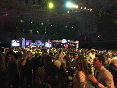 A large, diverse crowd of people mingling indoors under stage lights and screens. A banner reads 'Sundance Film Festival'.