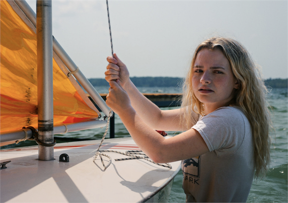 A blonde woman pulls on a rope attached to the mast of a small sailboat on a lake.