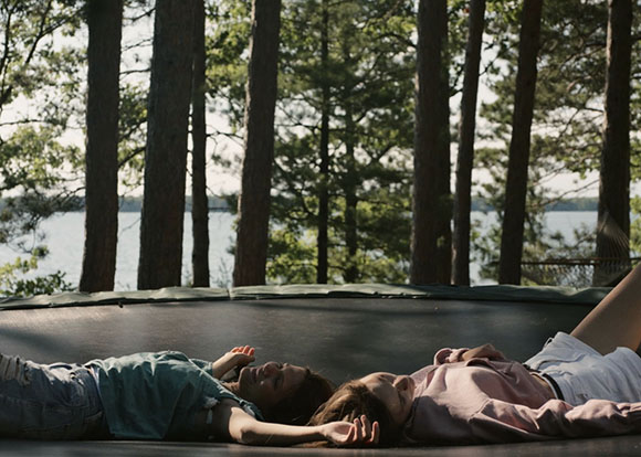Two people lie on a trampoline in a forest overlooking a large lake.