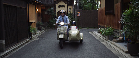 A person wearing a helmet rides a light-colored motor scooter down a narrow paved alleyway.