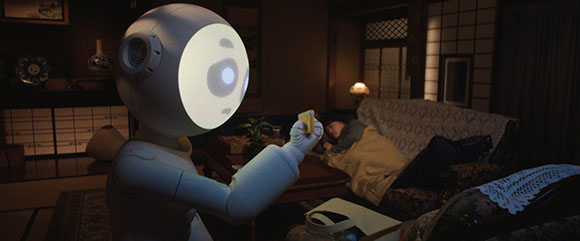 A white humanoid robot holds a small yellow object in a dimly lit living room near a person on a sofa.