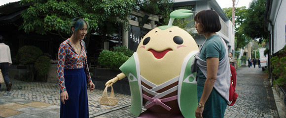 Two women stand on a stone path next to a large, smiling pear-shaped mascot wearing a green and pink outfit.