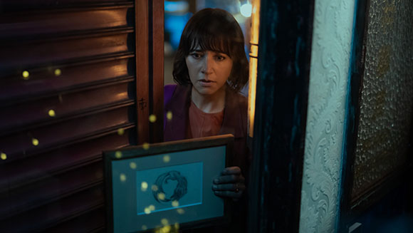 A woman with dark hair looks through a doorway while holding a framed picture.