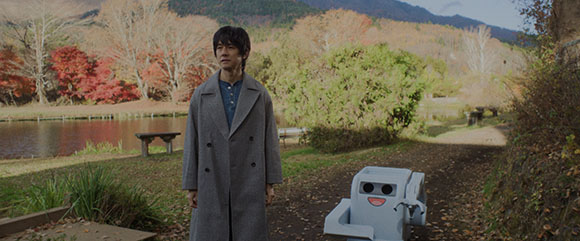 A man in a grey coat stands in a park next to a small, light blue robot near a pond.