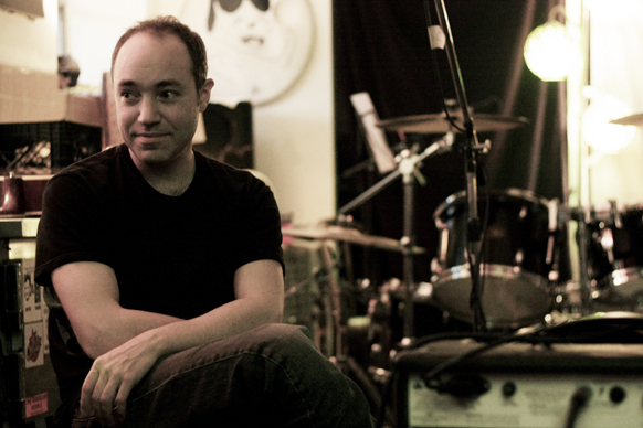 A man in a black t-shirt sits in a room filled with musical equipment and a drum set.