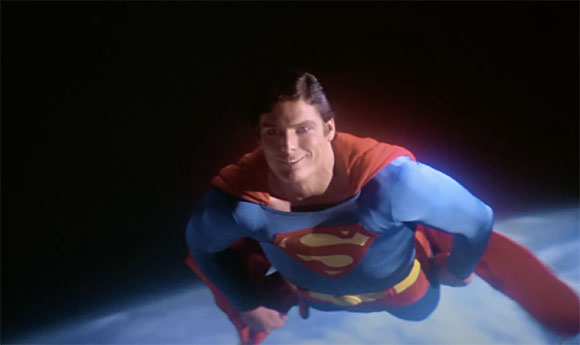 A man in a blue superhero suit with a red cape and 'S' emblem flies through a dark sky.
