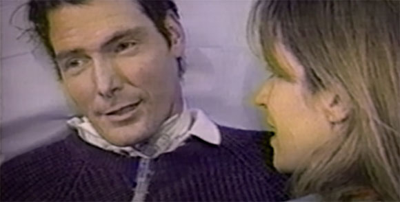 A man in a dark sweater looks toward a woman with light hair in profile.