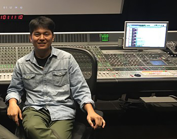 A man in a denim shirt sits in a chair before a large professional audio mixing console and computer monitor.