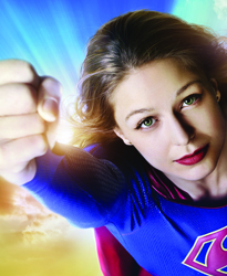 A woman in a blue superhero suit extends a clenched fist toward the camera against a bright, glowing background.